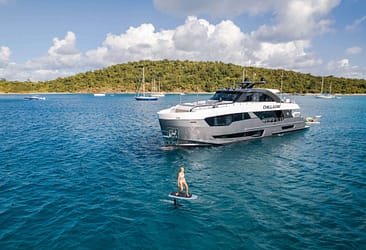 Luxury yacht on a serene blue sea with a person e-foiling nearby, surrounded by lush green islands and sailboats, showcasing a perfect day of adventure and relaxation on the water.