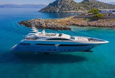 Luxury yacht anchored in clear blue waters near rocky coastline and mountains in the background.