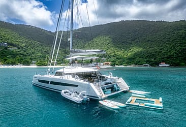 Luxury catamaran yacht anchored in tranquil turquoise waters with lush green hills in the background, featuring paddleboards and an inflatable dock.