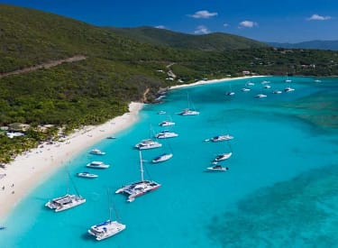 Aerial view of a tropical beach with white sand and turquoise waters, featuring anchored sailboats and a lush green hillside backdrop.