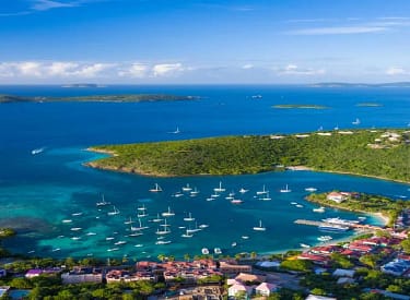 Aerial view of a vibrant coastal town with numerous boats in a tropical bay, lush green hills, and a cruise ship in the distance under a clear blue sky.