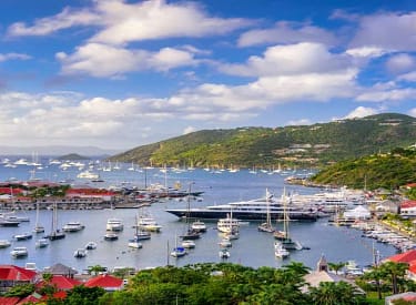 Panoramic view of Gustavia Harbor in Saint Barthélemy with moored yachts, red-roofed houses, lush green hills, and a partly cloudy sky.