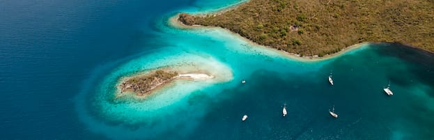Aerial view of a turquoise tropical coastline with small island and anchored sailboats in clear blue waters.