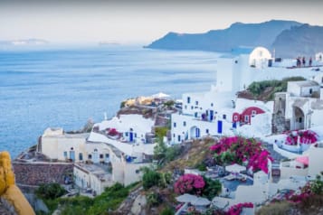 Scenic view of Santorini coastline with whitewashed buildings and vibrant bougainvillea, overlooking the Aegean Sea and distant islands.