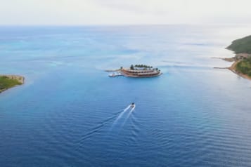 Aerial view of an idyllic island resort surrounded by vibrant blue ocean waters with a boat leaving a white trail, showcasing tropical getaway scenery.