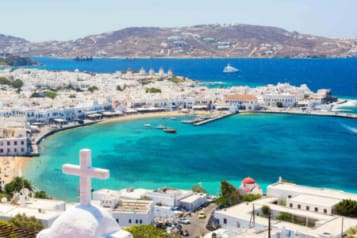 Aerial view of Mykonos town with whitewashed buildings, turquoise waters, and a coastline dotted with boats and windmills on a sunny day.