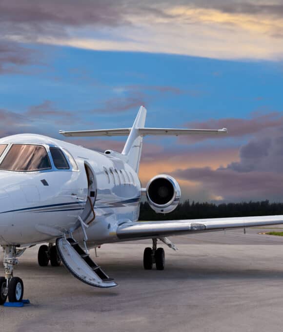 Private jet parked on an airport runway at sunset, with open door and step ladder, under a dramatic clouded sky.