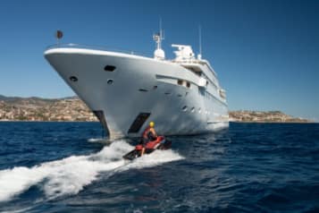 Luxury yacht sailing near the coast with person riding a jet ski in the foreground on a sunny day.