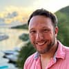 Smiling man in pink shirt by a scenic waterfront at sunset, with blurred greenery and boats in the background.