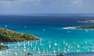 View of a scenic Caribbean bay with numerous sailboats on turquoise waters, surrounded by lush green islands under a partly cloudy sky.