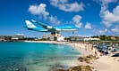 KLM airplane flying low over Maho Beach in Saint Martin, with tourists watching from the shore and a clear blue sky above.