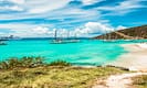 Tropical beach with turquoise water, sailboats anchored near the shore, and a hillside landscape under a blue sky.