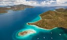 Aerial view of tropical islands with lush green hills and turquoise waters, surrounded by boats and expansive ocean, under a partly cloudy sky.