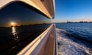 Luxury yacht cruising at sunset with city skyline and ocean waves, reflecting the vibrant evening sky.