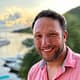 Smiling man in pink shirt by a scenic waterfront at sunset, with blurred greenery and boats in the background.