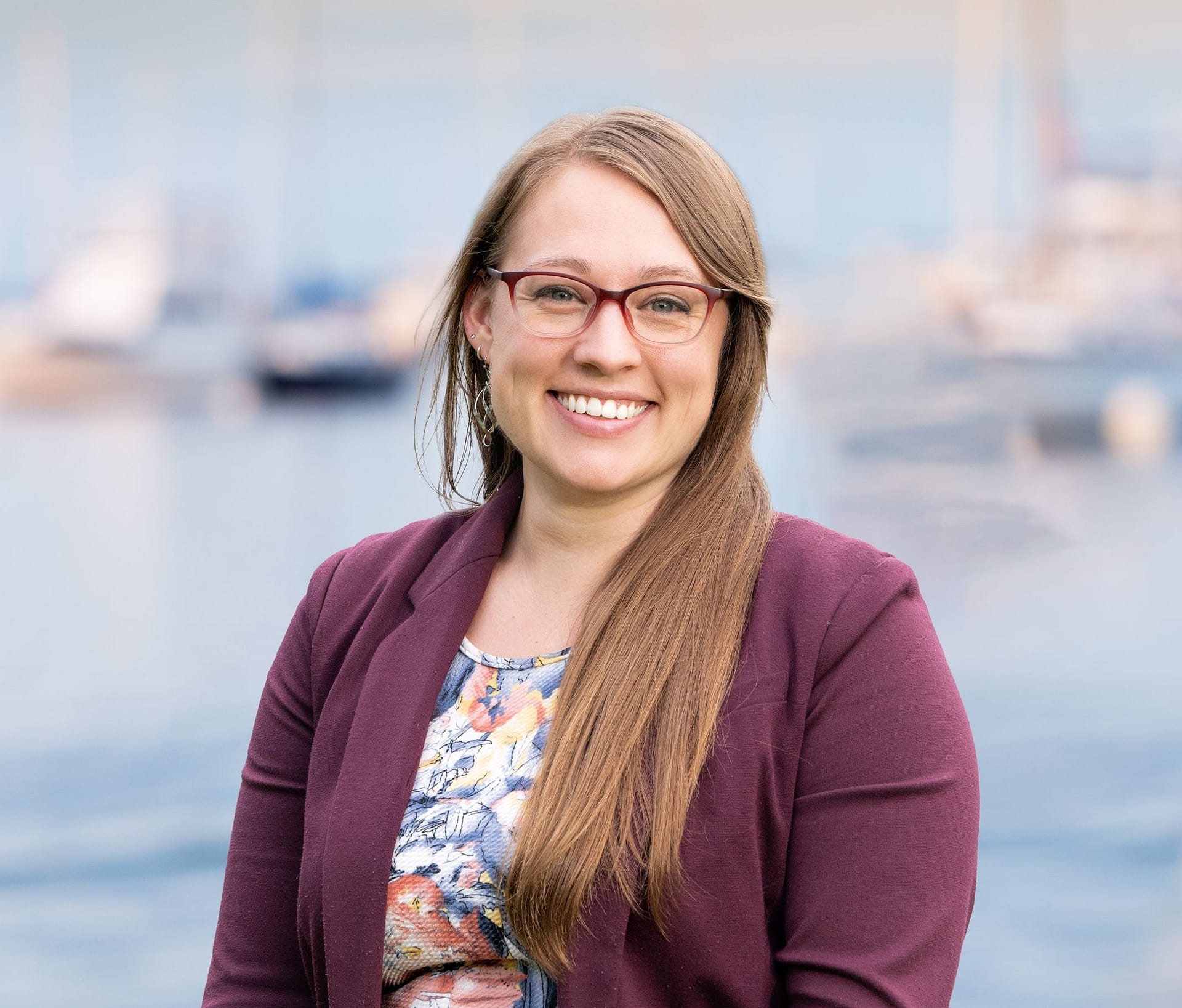 Smiling woman with long hair wearing glasses and a purple blazer, standing in front of a blurred marina background.
